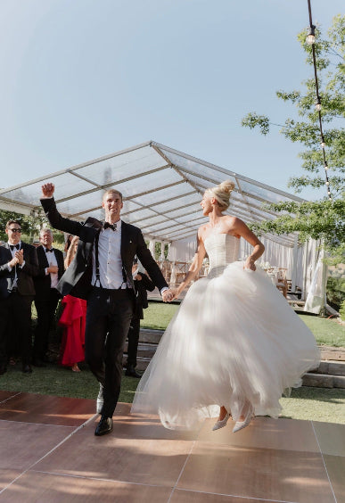 A bride in a strapless white tulle wedding gown and a groom in a black tuxedo dance together on an outdoor patio under a clear sky, with guests and a marquee tent in the background.